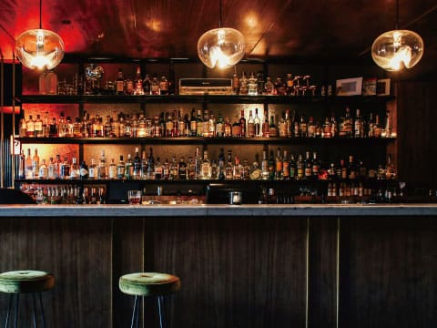 Rows of spirit bottles lining a bar's back shelves, illuminated by warm pendant lighting above a wooden counter