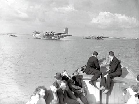 A black-and-white 1938 photograph of passengers on a boat approaching seaplanes moored at Foynes Airport in Ireland