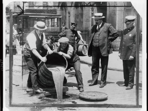 A black-and-white historical photo of Prohibition-era officials pouring confiscated bootleg alcohol into a street drain while police officers look on