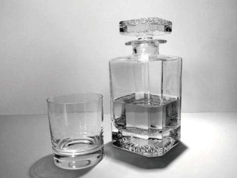 A black and white photo of a crystal decanter filled with clear liquid beside an empty rocks glass