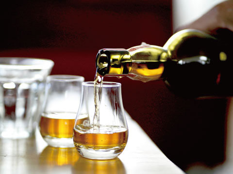 Whisky is poured from a bottle into a row of tulip-shaped tasting glasses on a bar counter.