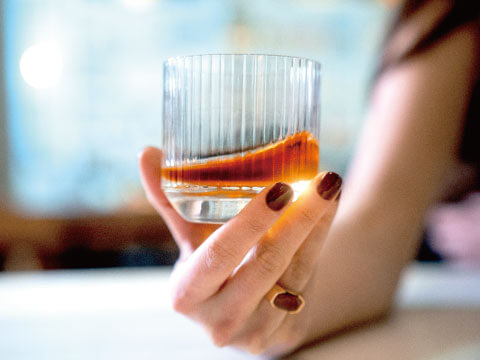 A woman's hand holds a ridged rocks glass containing whiskey on the rocks near a sunlit window.