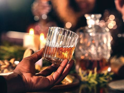 A hand holds a ridged rocks glass of whiskey with candles and a crystal decanter in the softly lit background.