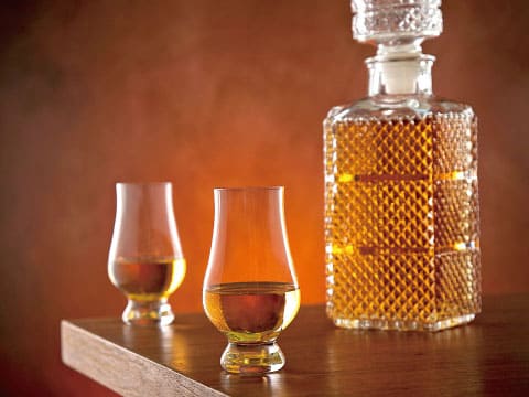 Two tulip-shaped Glencairn glasses of whisky sit on a wooden table beside a cut-crystal decanter.