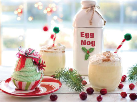 Two glasses of eggnog topped with whipped cream and cinnamon, garnished with candy cane stirrers, next to a jug labeled Egg Nog and fresh cranberries