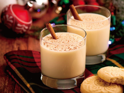 Two glasses of eggnog garnished with cinnamon sticks and a dusting of nutmeg, served on a plaid cloth with shortbread cookies