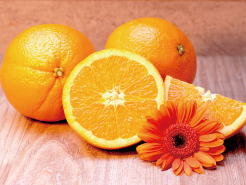 Whole oranges beside a sliced orange half and wedge, garnished with an orange gerbera daisy on a wooden surface