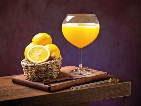 A glass of fresh orange juice beside a basket of whole and halved oranges on a wooden cutting board