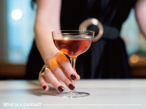 A woman in a black dress picking up a short cocktail in a coupe glass from a marble bar counter