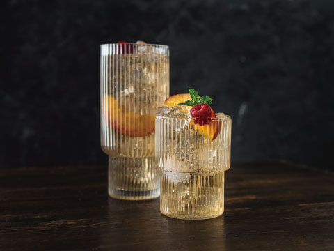 Two amber-colored cocktails served over crushed ice in ribbed glasses, garnished with peach slices and mint, illustrating how ice machines and distillation shaped modern cocktails