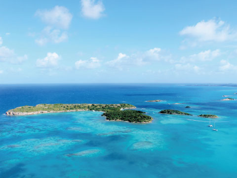 Aerial view of a small green Caribbean island surrounded by turquoise water and coral reefs under a blue sky