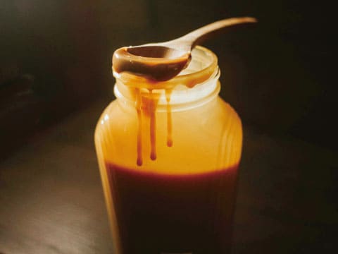 Rich caramel syrup drips from a wooden spoon into a glass jar.