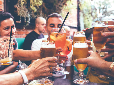 A group of friends clinking together beer glasses, a cocktail coupe, and other assorted glassware in a toast at an outdoor table