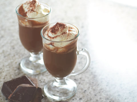 Two footed glass mugs of Irish coffee topped with whipped cream and dusted with cocoa powder, served beside chunks of dark chocolate