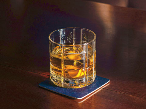 An Old Fashioned glass with a cut crystal pattern, filled with whiskey and a single large ice cube, set on a coaster