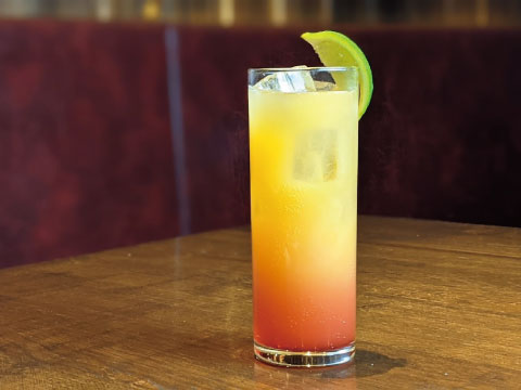 A tall Collins glass filled with a layered orange-to-red tropical cocktail, garnished with a lime wheel and served over ice