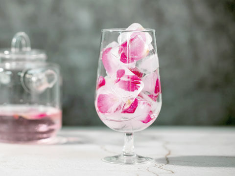 A footed goblet glass filled with pink flower-infused ice cubes, with a decanter of rose-colored liquid in the background