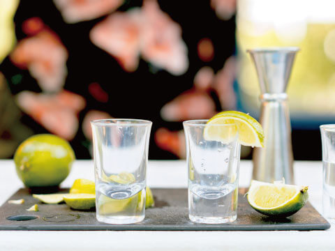 Two clear shot glasses garnished with lime wedges, served alongside fresh limes and a stainless steel jigger on a serving tray