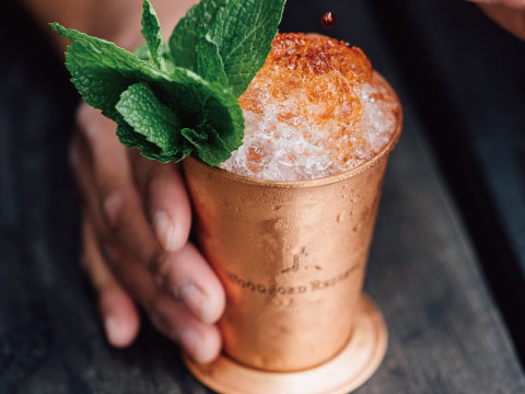 A metal Julep cup filled with crushed ice and a mint sprig garnish, held by hand on a wooden bar counter