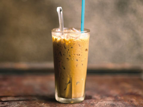 A tumbler glass filled with iced coffee and crushed ice, served with a metal spoon and a blue straw