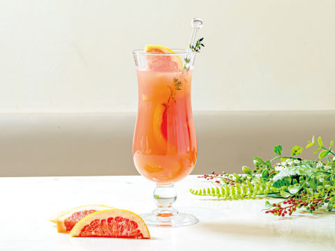 A Hurricane glass filled with a pink tropical cocktail, garnished with grapefruit slices and fresh herbs, with citrus wedges alongside