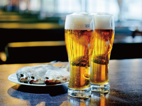 Two tall, ribbed Pilsner glasses filled with golden beer and foam, served alongside a plate of grilled whole fish