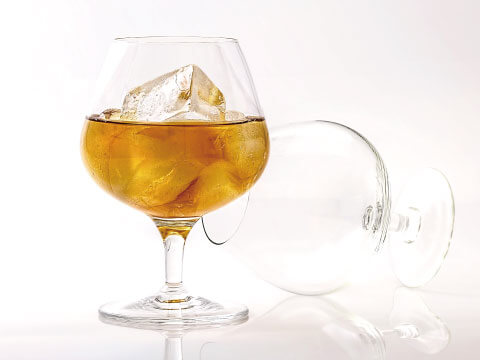 A wide-bowled brandy snifter filled with brandy and a large ice cube, with an empty glass tipped on its side beside it