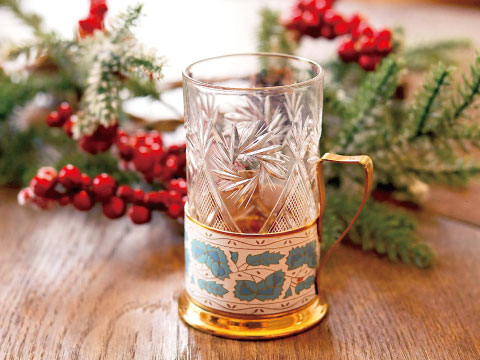 A cut-crystal hot drink glass set in a decorative gold metal holder with a handle, surrounded by holiday berries and pine branches