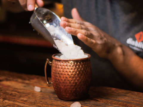 A hammered copper Moscow Mule mug being filled with crushed ice using a metal scoop, on a wooden bar counter