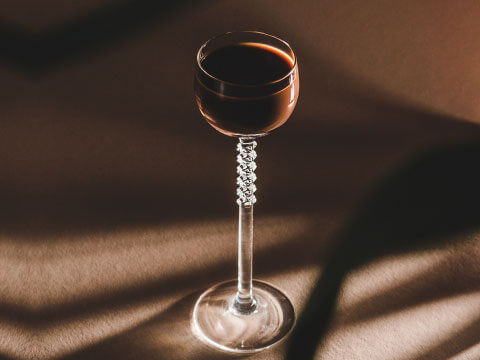A small liqueur glass with a long, decorative beaded stem, filled with a dark reddish-brown liqueur