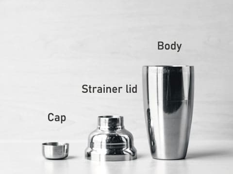 A disassembled three-piece cobbler shaker labeled with its parts: the body, strainer lid, and cap, arranged from largest to smallest
