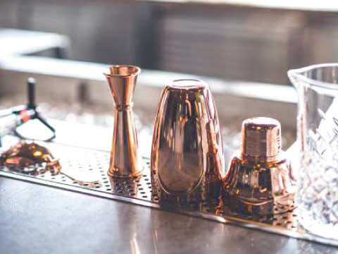 A copper three-piece cocktail shaker disassembled into its body, strainer top, and cap, displayed on a bar mat beside a jigger and mixing glass