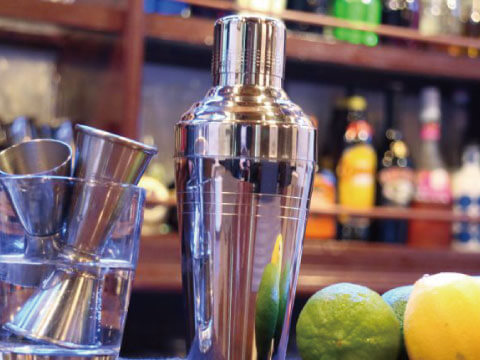 A stainless steel professional cocktail shaker on a bar counter beside jiggers and fresh limes and lemons, with bottles in the background
