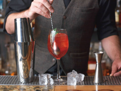 A large Boston shaker tin beside a red cocktail in a wine glass, garnished with mint, with ice cubes and a jigger on the bar