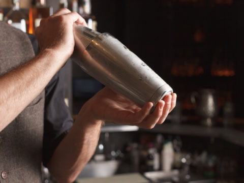 A bartender gripping a metal Boston shaker with both hands, ready to shake, in a dimly lit bar