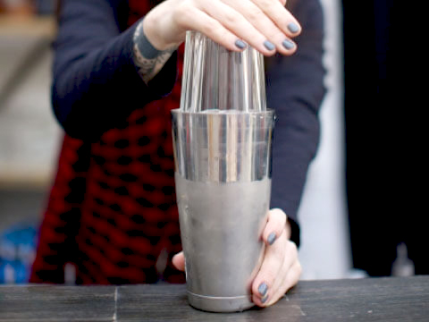 Hands fitting a pint glass into a metal tin to assemble a Boston shaker on a bar counter