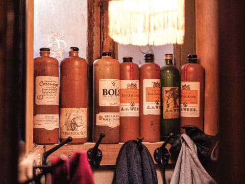 A row of traditional stoneware and glass Genever bottles, including Bols and other classic Dutch brands, displayed on a wooden shelf