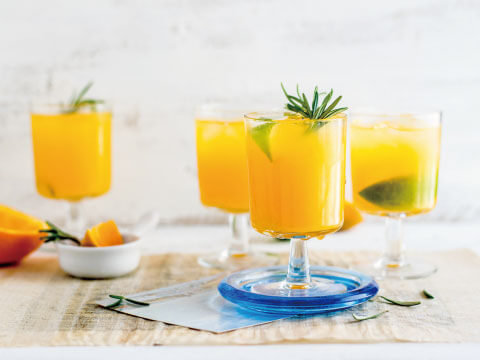 Several glasses of orange-colored cocktails garnished with rosemary and lime wedges, arranged on a woven table runner
