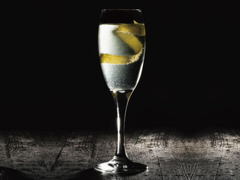 A Crusta-style cocktail in a champagne flute, lined inside with a long spiral of lemon peel, served on a dark textured background