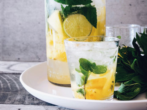 A citrus Cooler-style cocktail with pineapple, lime, and mint served in a glass and pitcher, garnished with fresh mint leaves