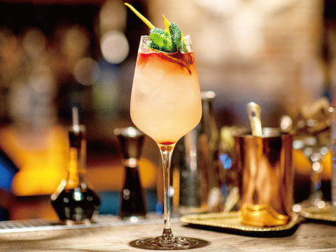 A pale pink Sour-style cocktail served in a stemmed glass with a red wine float, garnished with fresh mint and a straw