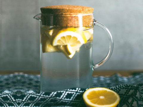A glass pitcher of lemon-infused water with a cork lid, garnished with lemon slices, set on a patterned cloth