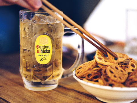 A Suntory Whisky highball served in a mug with ice at a Japanese izakaya, paired with fried lotus root chips eaten with chopsticks