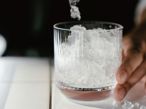 Crushed ice being poured into a ridged rocks glass, the base of a Fix-style cocktail, held by hand on a marble counter