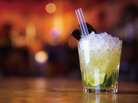 A green Frappé-style cocktail piled high with crushed ice in a rocks glass, garnished with lime and served with striped straws