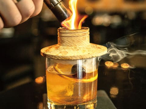 A torch ignites the wooden lid of a smoking cloche covering a whiskey cocktail with orange peel and a large ice cube.