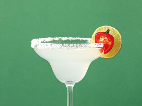 A margarita glass with a salted rim is garnished with a lime wheel and a small red chili pepper.