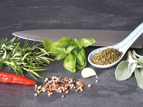 Rosemary, basil, sage, chili, garlic, and peppercorns are arranged on a dark surface beside a chef's knife and a spoonful of dried herbs.