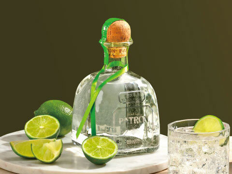 A bottle of Patrón Silver tequila surrounded by fresh limes, with a tequila cocktail over ice in the background