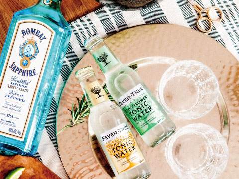 A bottle of gin sits beside tonic water bottles and empty glasses arranged on a copper tray.